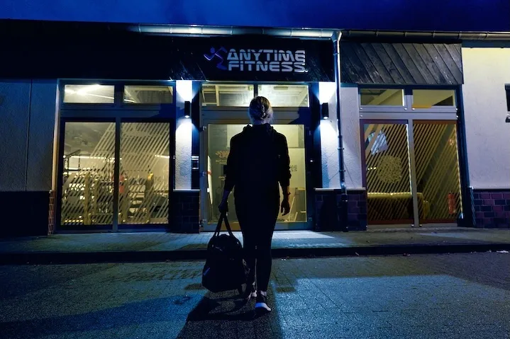 Rückansicht einer Frau mit Sporttasche, die nachts auf den hell beleuchteten Eingang eines Anytime Fitness Studios zugeht.
