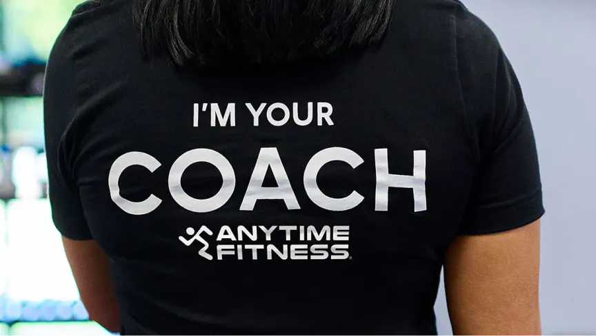 Rückansicht einer Person in einem schwarzen T-Shirt mit dem Aufdruck: 'I'M YOUR COACH' und dem 'ANYTIME FITNESS'-Logo darunter.