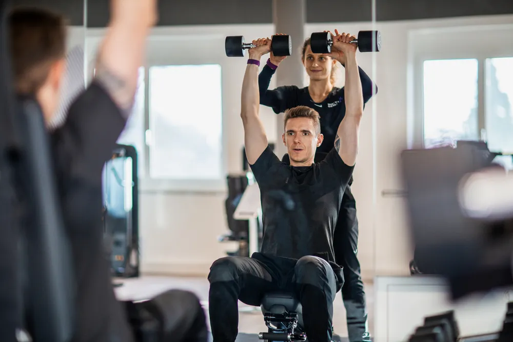 Man lifting dumbbells overhead while seated in a gym, with a trainer assisting him from behind.