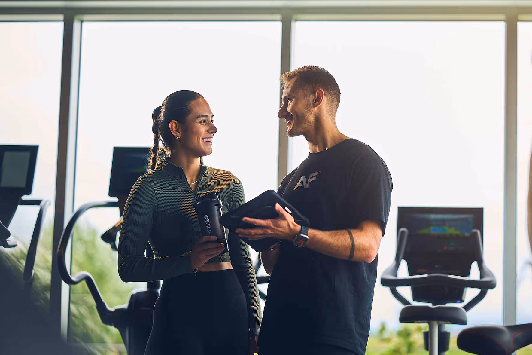 Des entraîneurs de conditionnement physique féminins et masculins parlant au gymnase avec des vélos d'exercice et de grandes fenêtres derrière eux.