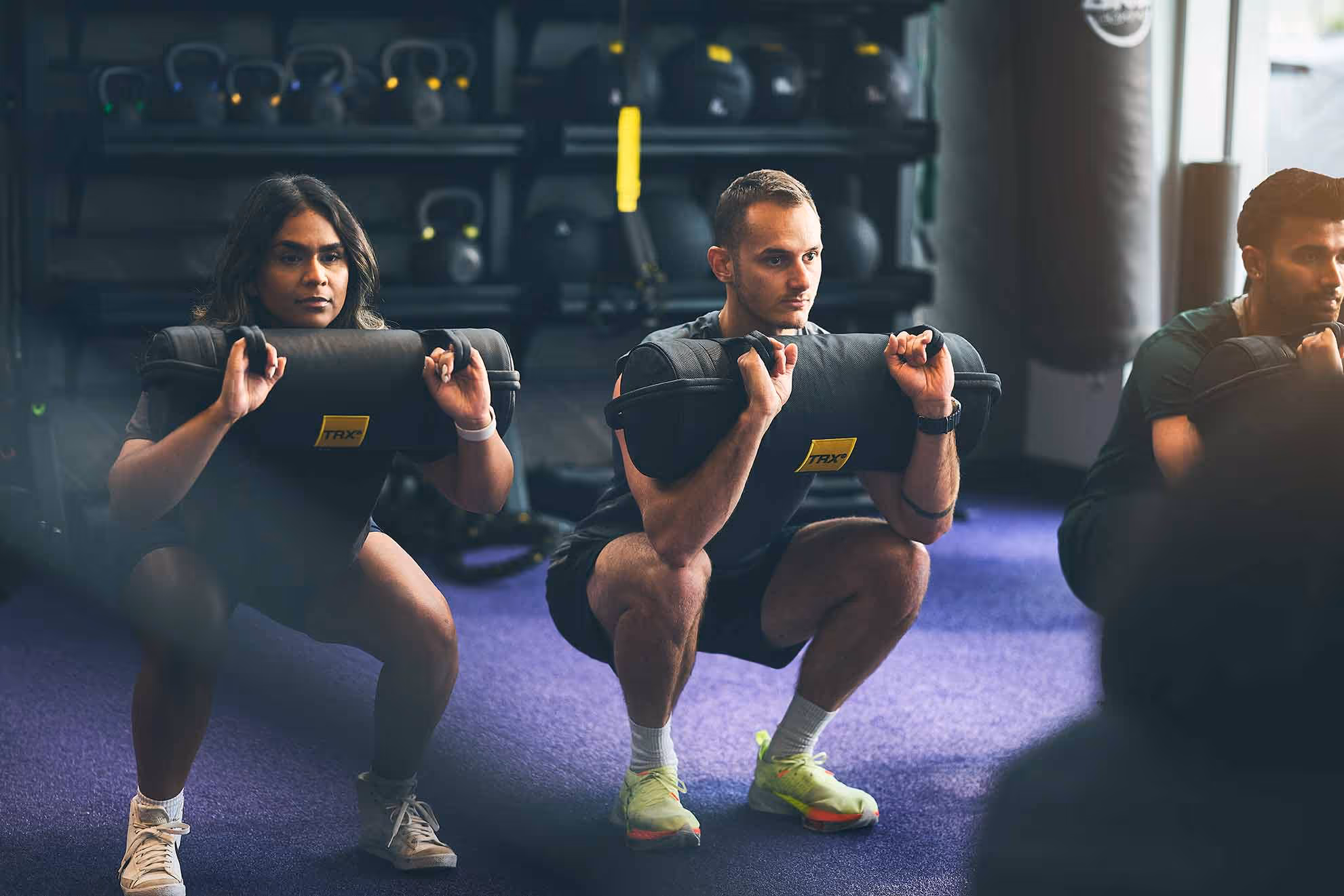 Trois personnes dans un gymnase faisant des squats létés tenant de grands sacs de sable noirs sur les épaules.