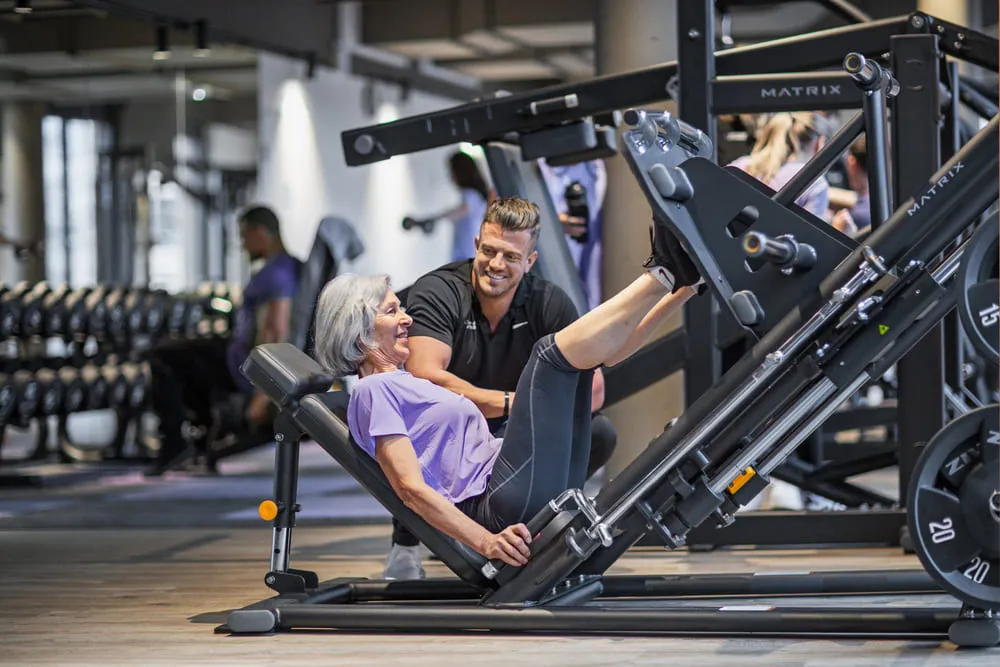 Older woman exercises leg press machine at gym, assisted by personal trainer, with other gym-goers and equipment visible in background.
