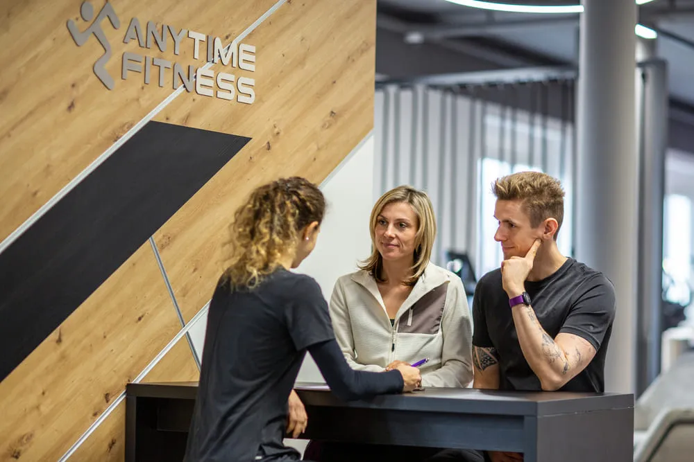 Three people at an Anytime Fitness front desk, with two listening and one speaking while holding a pen.
