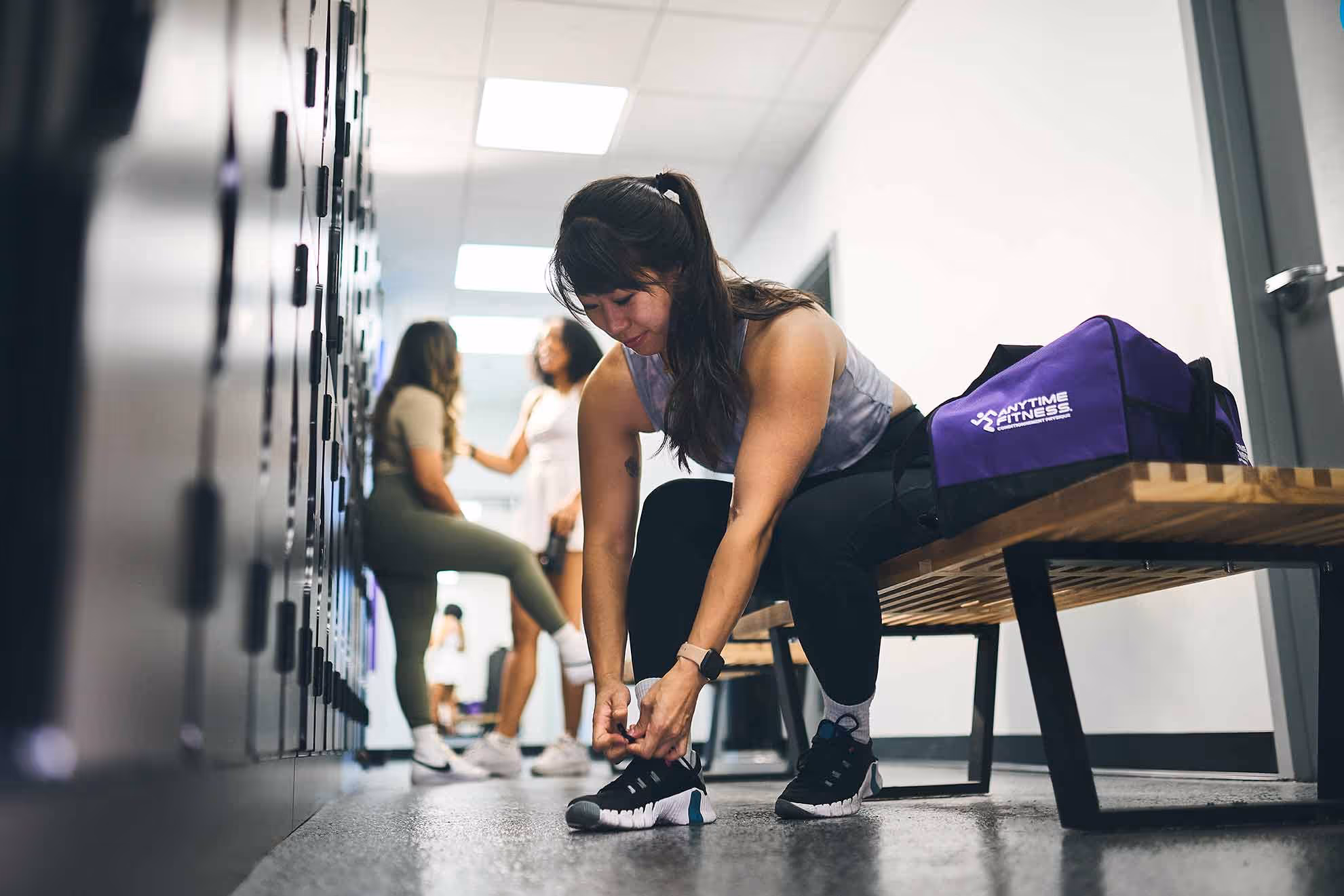 Femme attachant des lacets sur un banc dans les vestiaires du gymnase avec d'autres personnes et un sac de sport à proximité.
