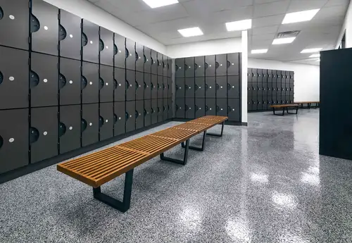 Modern gym locker room with black lockers, wooden benches, and bright ceiling lights.