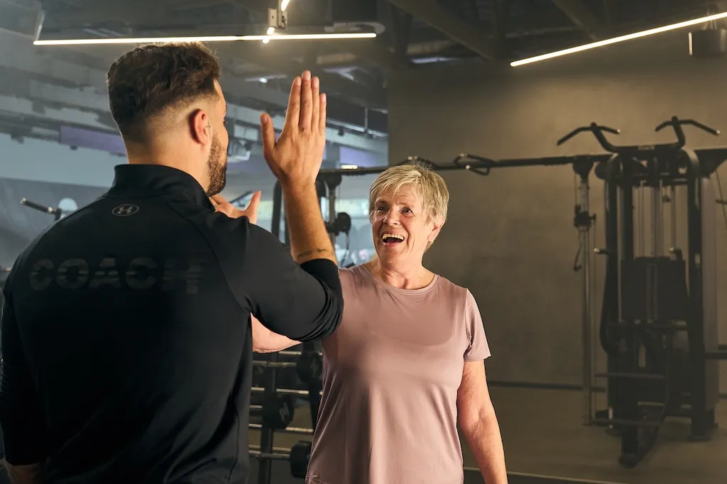 Eine Anytime Fitness Trainerin unterstützt ein Mitglied bei Kniebeugen mit der Langhantel im Trainingskäfig.