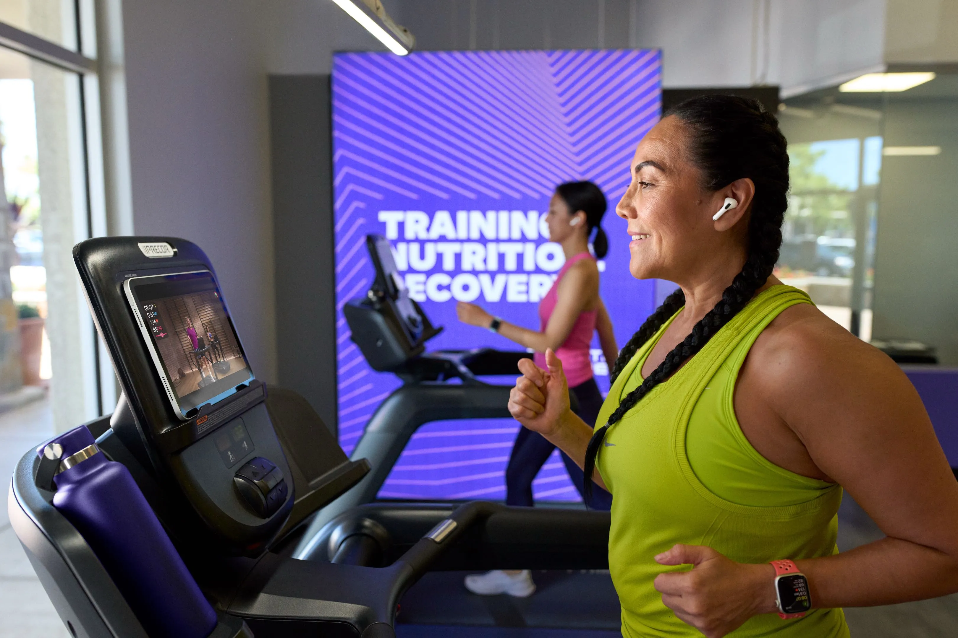 Two women running on treadmills indoors, one watching fitness video, with training advice on screen.