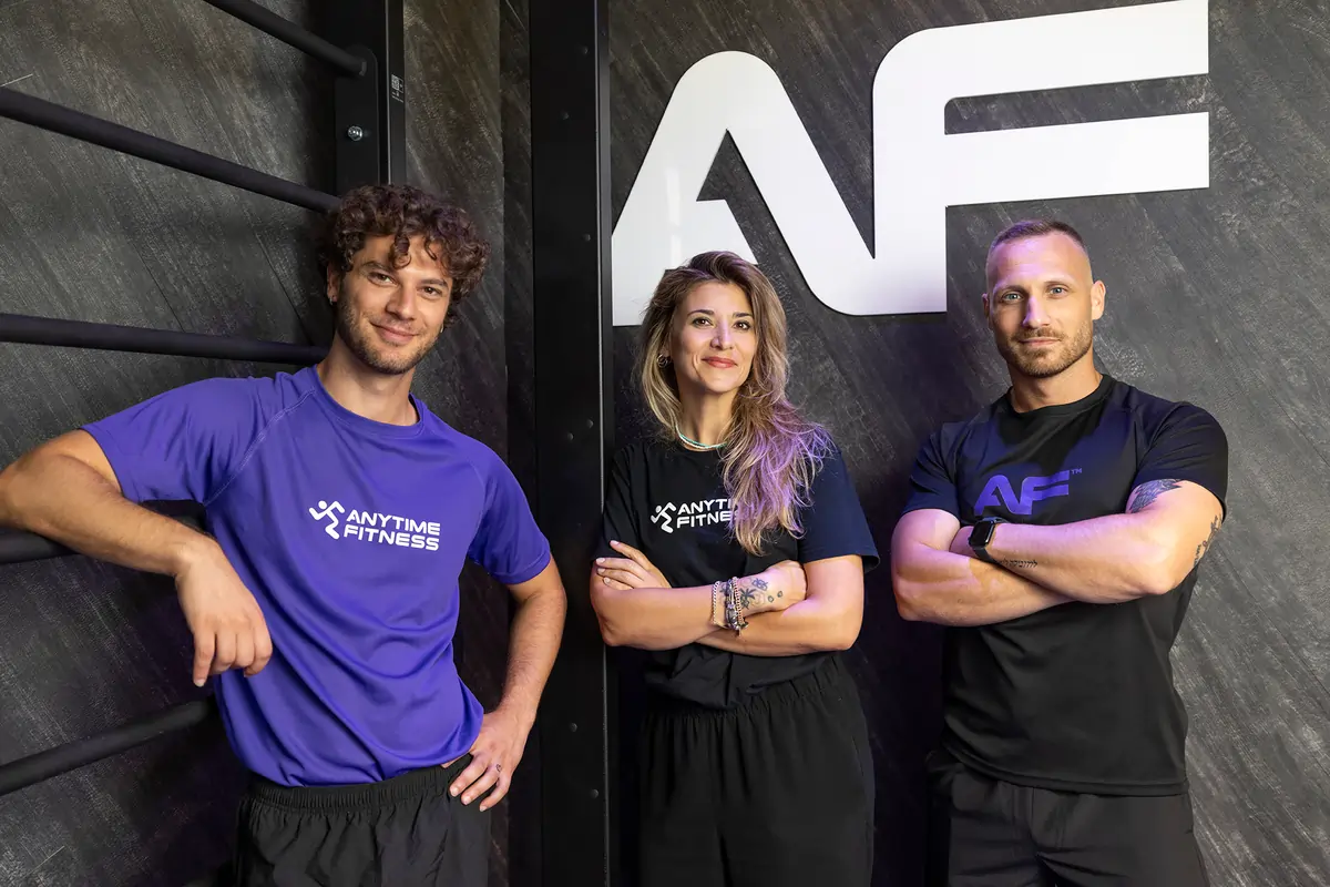 Tre istruttori di fitness in piedi accanto a un muro con il logo Anytime Fitness, sorridono alla telecamera.