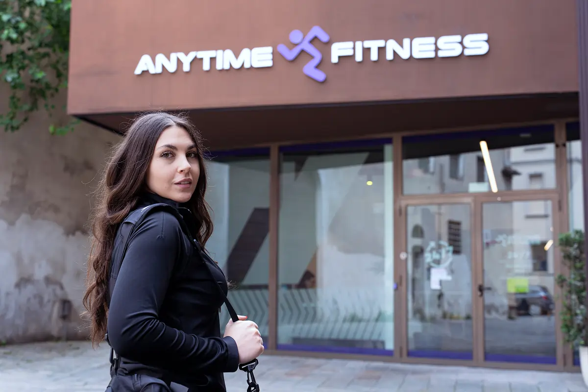 Una donna con i capelli lunghi in giacca nera si trova davanti all'ingresso di una palestra Anytime Fitness.