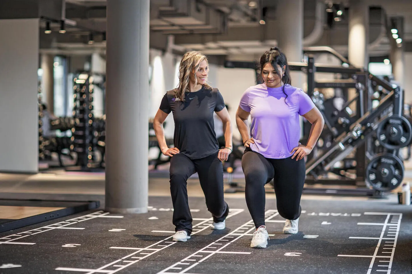Two women in athletic wear performing lunges in a gym with weightlifting equipment in the background.