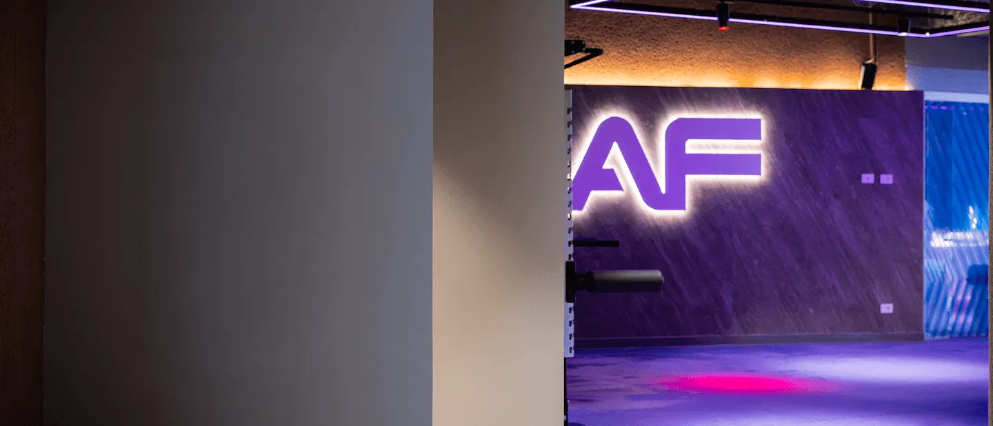 Interno di una moderna palestra Anytime Fitness con il logo monumentale AF illuminato a LED viola su una parete scura texturizzata.