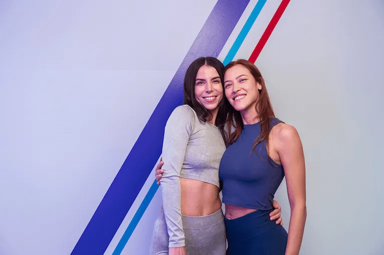 Due donne sorridenti in abbigliamento da fitness posano insieme all'interno di una palestra Anytime Fitness, davanti a una parete con strisce diagonali colorate.