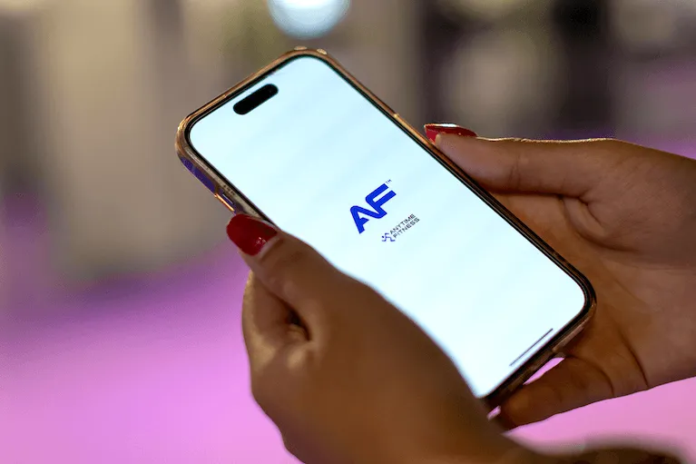 Mani con unghie rosse che reggono uno smartphone con il logo Anytime Fitness sullo schermo.