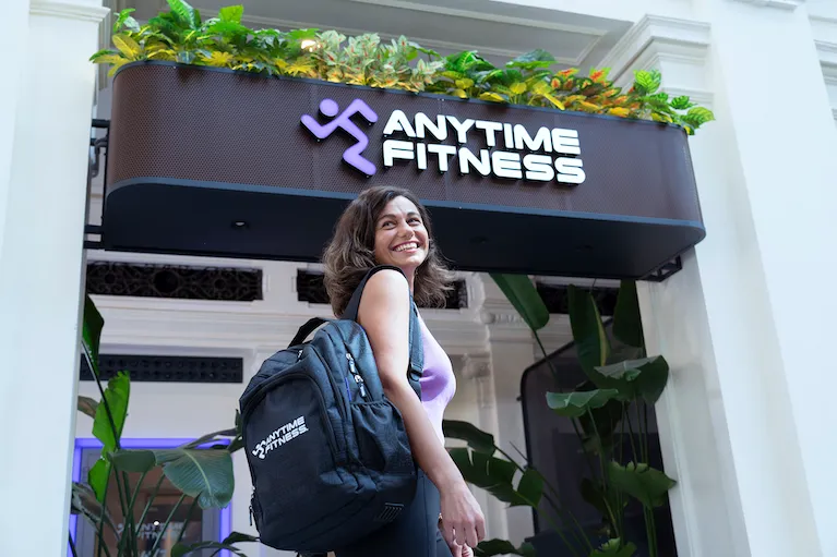 Una giovane donna sorridente con uno zaino Anytime Fitness cammina davanti all'ingresso di una palestra, sotto l'insegna luminosa del club decorata con piante.