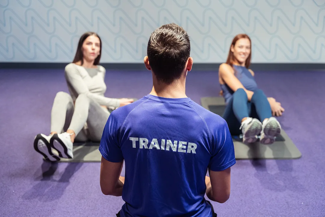 Un personal trainer in maglietta blu segue da dietro due donne che eseguono esercizi per addominali sui tappetini in una sala fitness con pavimento viola.