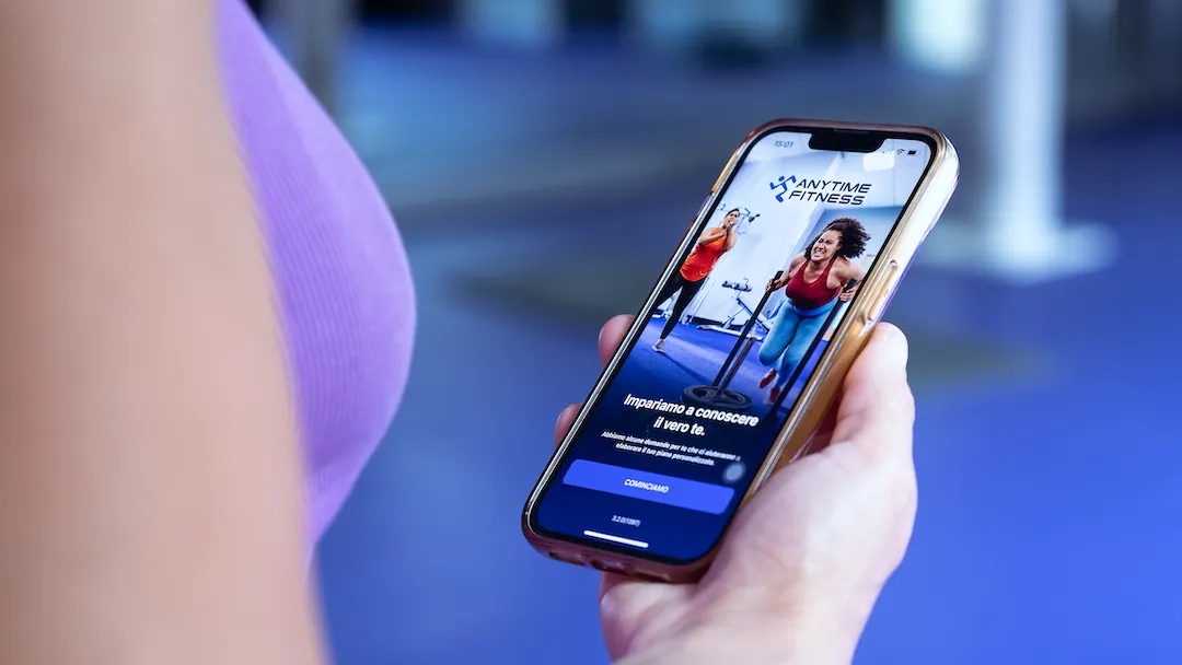 Una persona tiene in mano uno smartphone con l'applicazione di Anytime Fitness aperta sullo schermo all'interno di una palestra.