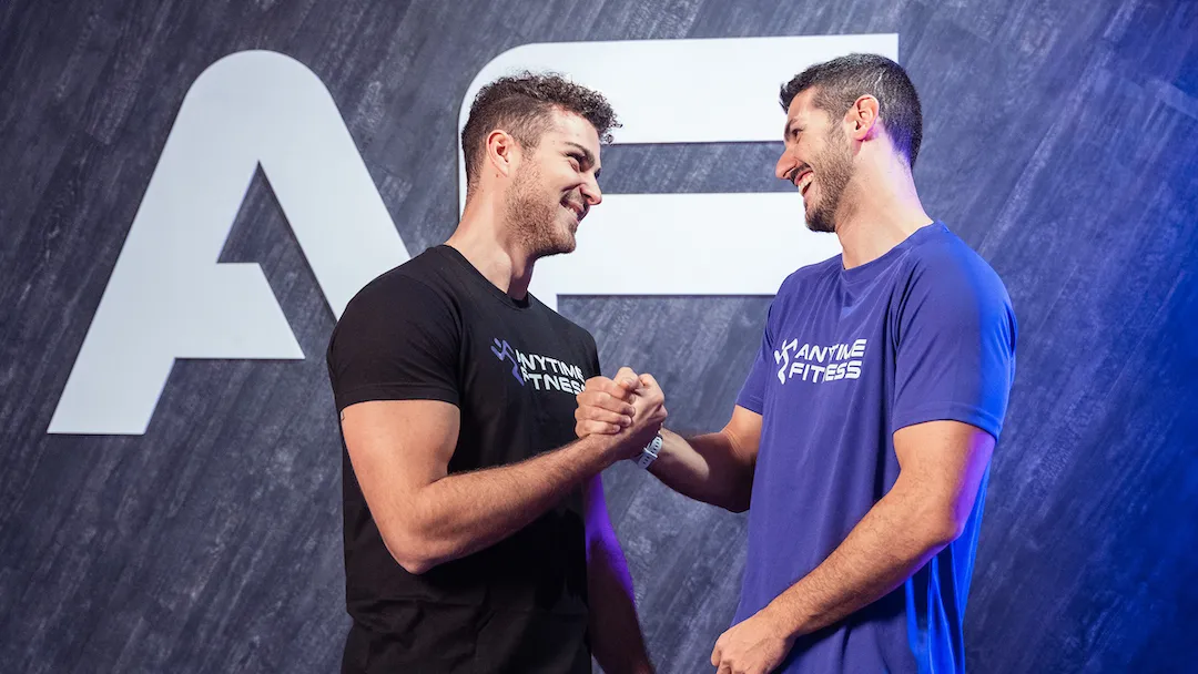 Due personal trainer con magliette Anytime Fitness si stringono la mano e sorridono davanti al grande logo AF.