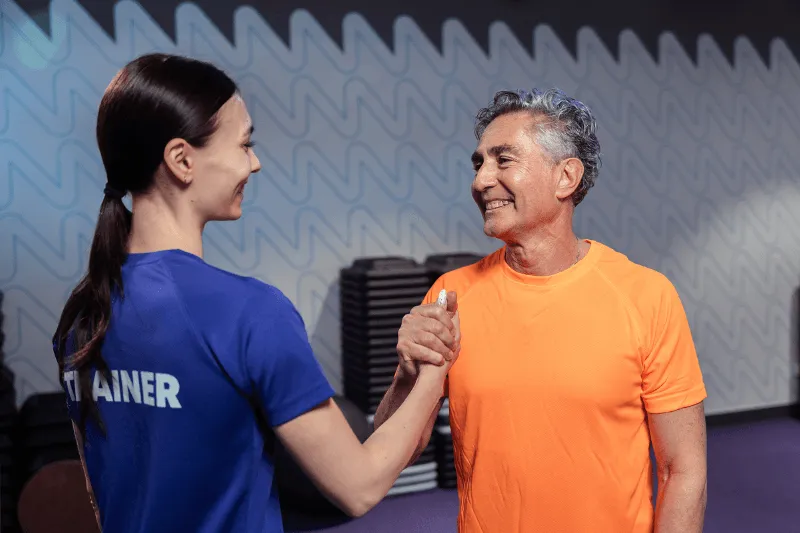 Una giovane donna sorridente in abbigliamento sportivo si appoggia a una struttura in palestra mentre controlla il suo smartphone, circondata da luci soffuse viola tipiche del club Anytime Fitness.