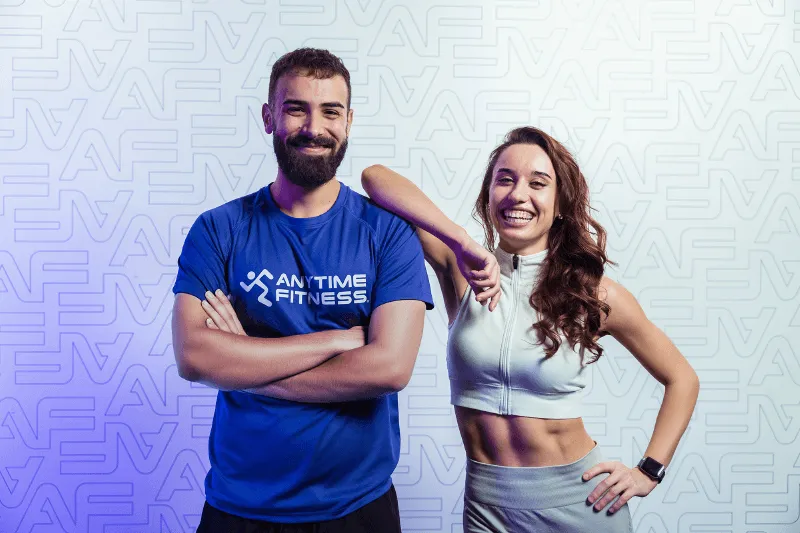 Una donna felice e rilassata siede su una panca da fitness tenendo in mano una borraccia viola, all'interno di un club Anytime Fitness luminoso e moderno.