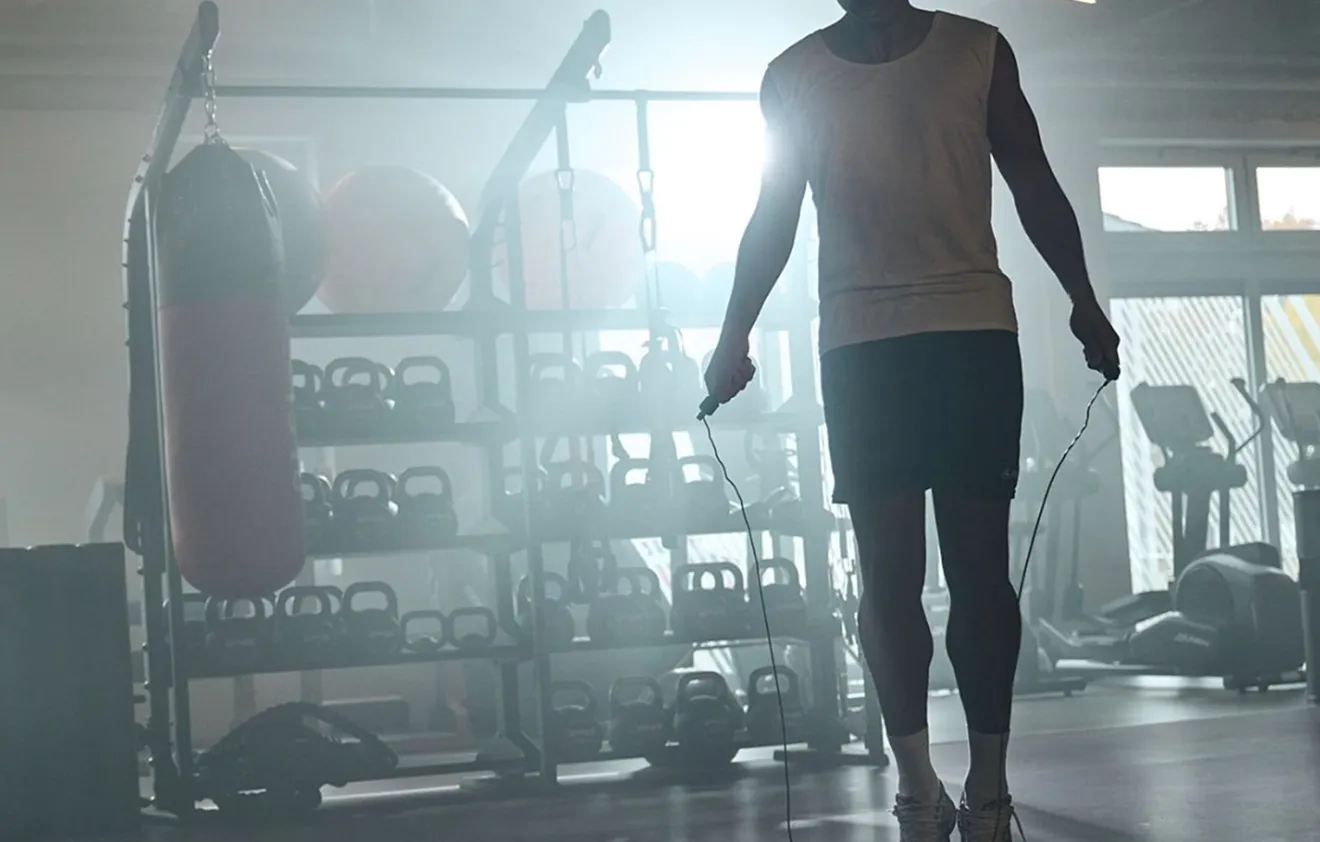 Person beim Seilspringen im Fitnessstudio mit Gewichten, Kettlebells und Boxsack im Hintergrund.