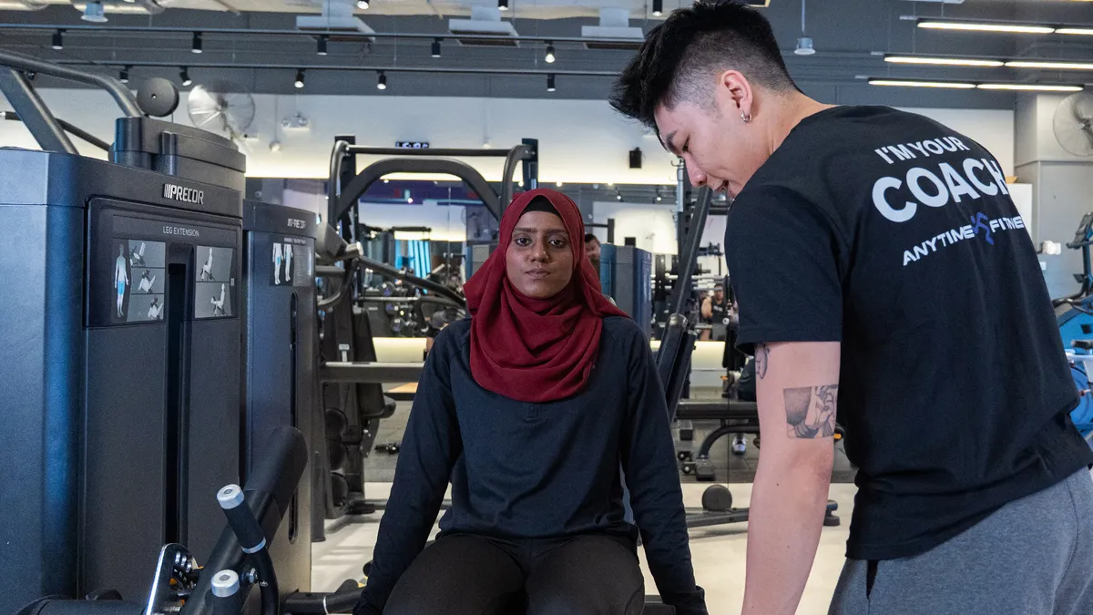 Woman in a red hijab using gym leg extension machine while coach guides her workout.