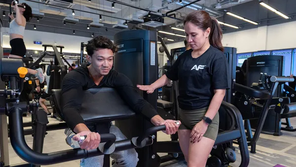 Trainer assists man using arm curl machine in a gym with exercise equipment in background.