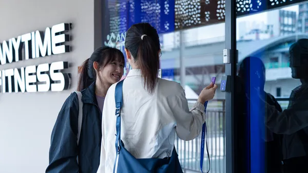 Two women at a gym entrance, one scanning a card to enter while the other smiles.