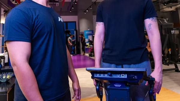 Two men standing near exercise equipment in a gym setting with focus on their torsos and arms.