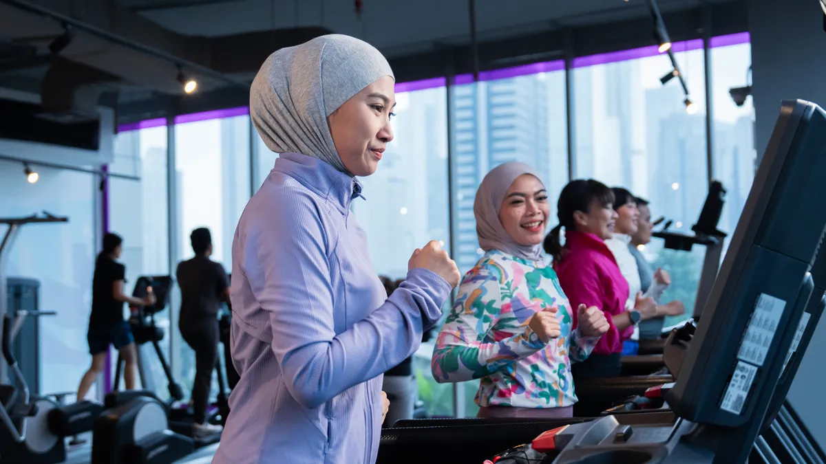 Women, some wearing hijabs, exercising on treadmills in a gym with city buildings outside.