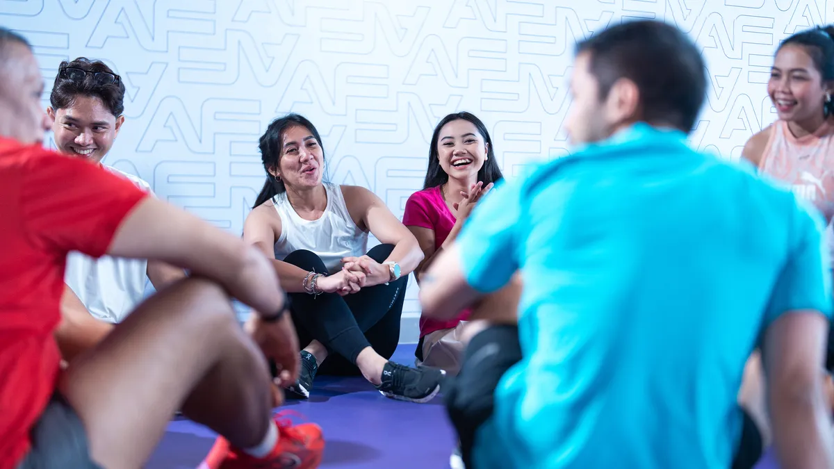 Group of six young adults sitting on a purple gym floor, smiling and chatting in workout clothes.
