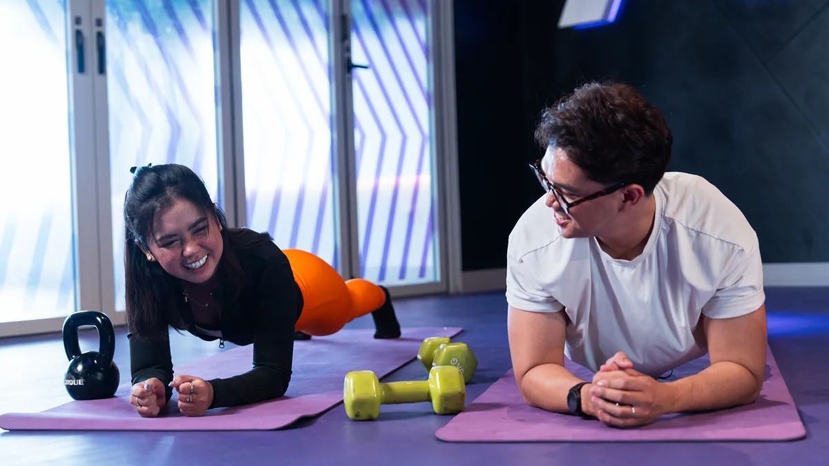 Smiling man and woman doing plank exercise facing each other on purple mats with weights nearby.