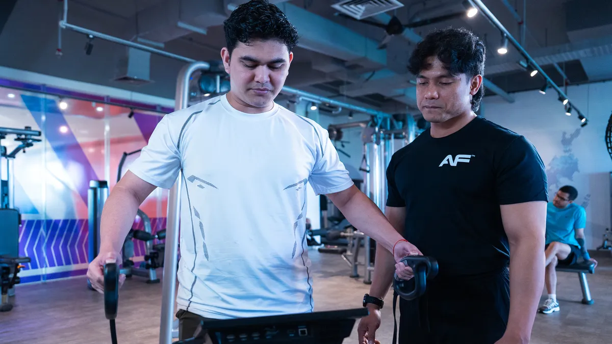 Two men at a gym, one using fitness equipment and another instructing him.