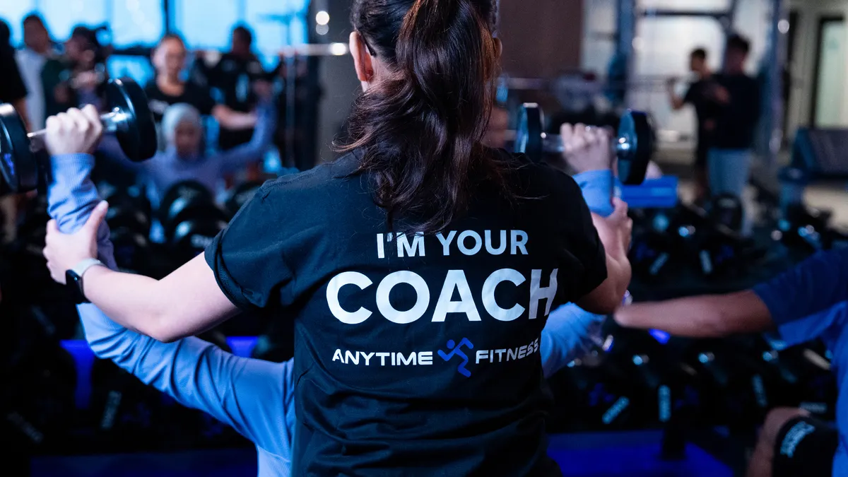 Fitness coach assisting a person lifting dumbbells in a gym with blurred background.