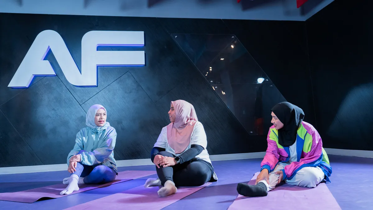 Three women in hijabs sitting on purple mats stretching legs in a gym with AF logo on the wall.