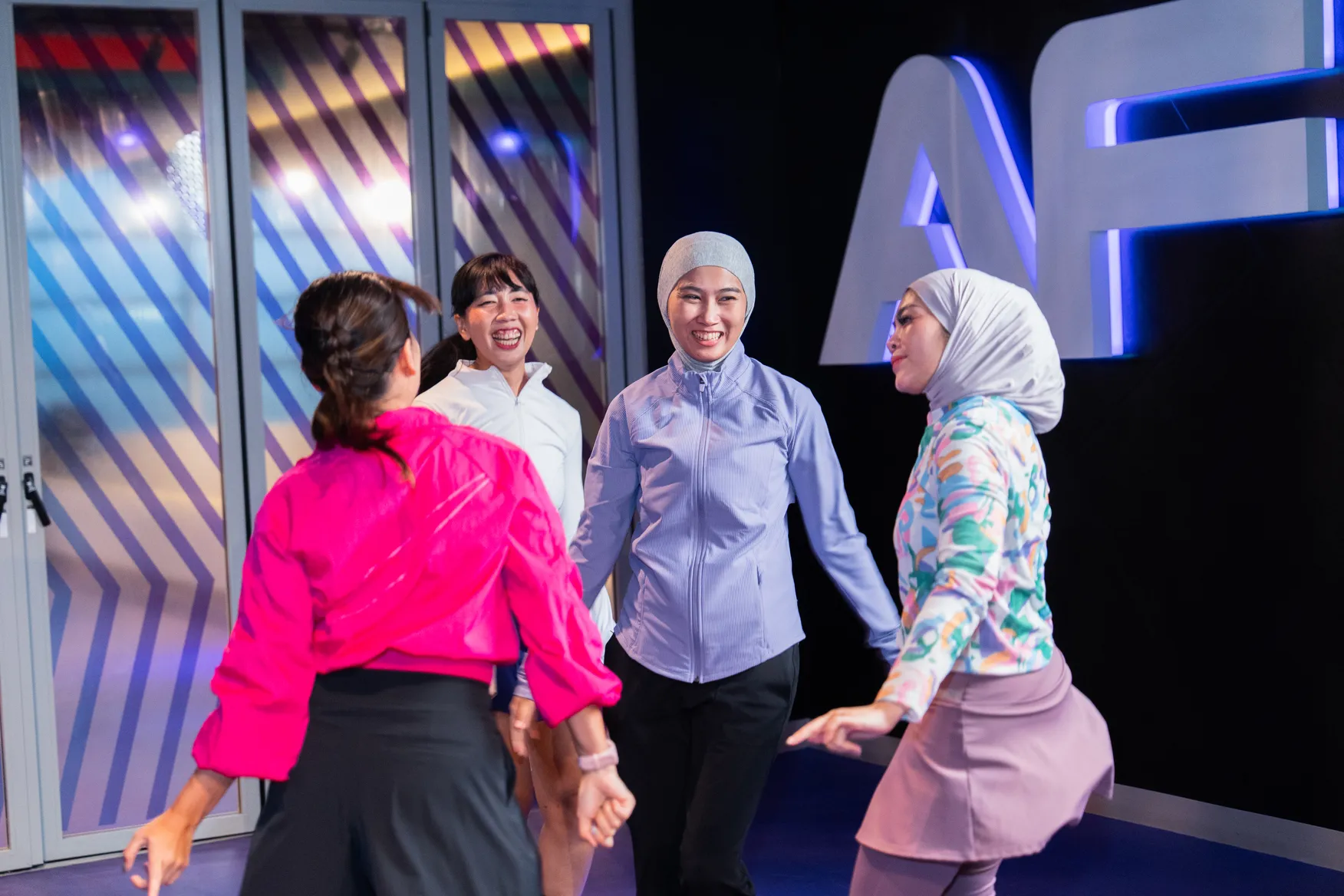 Four women holding hands and smiling in a brightly lit indoor space with colorful decor.