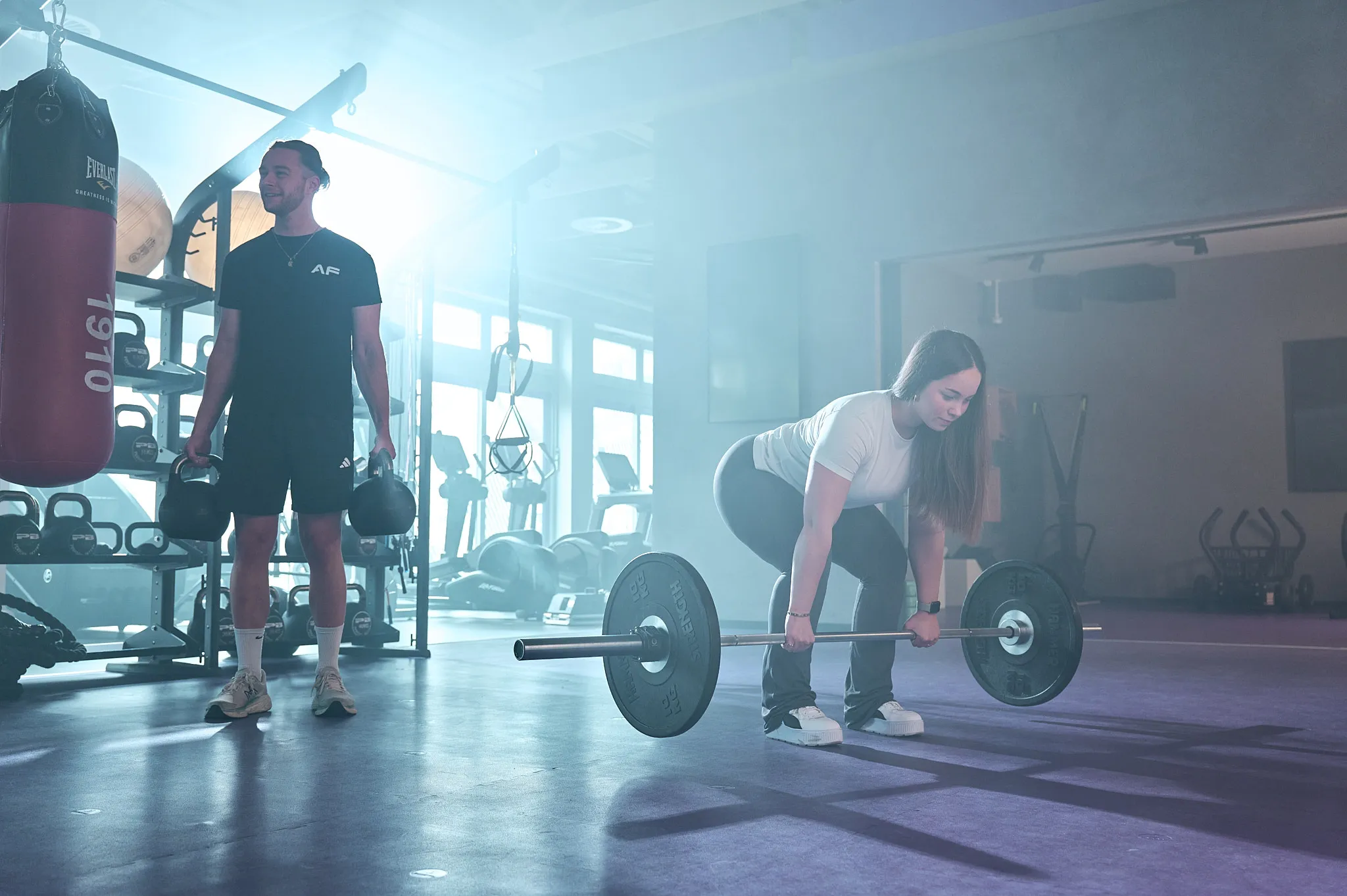 Eine Frau beim Heben einer Langhantel und ein Mann mit Kugelhanteln in einem Fitnessstudio mit Trainingsgeräten.