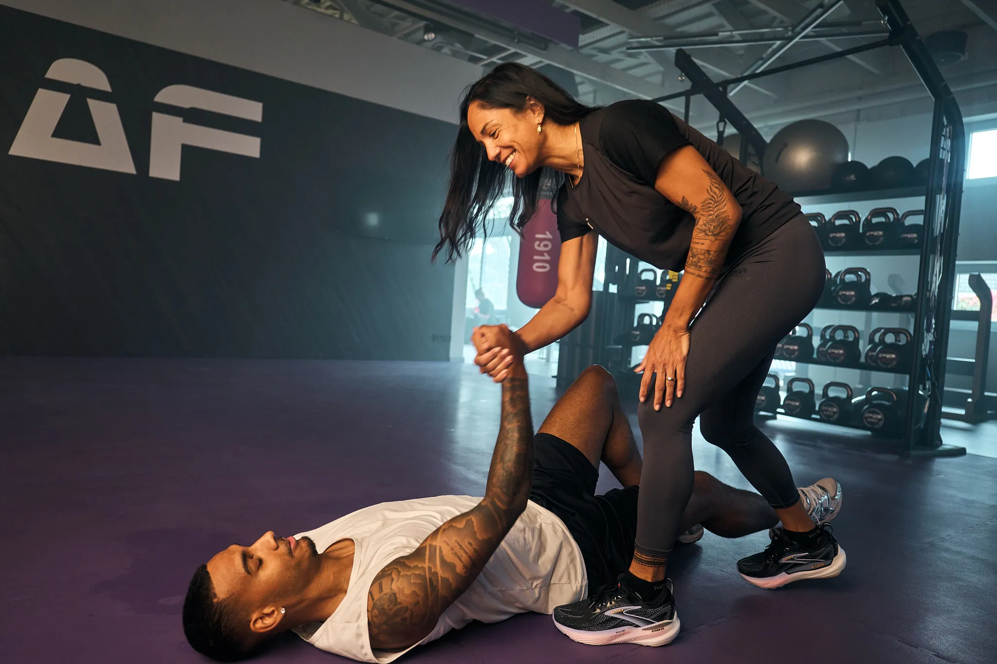 Fitte Frau hilft Mann beim Tätowieren vom Boden des Fitnessstudios, lächelt und hält seine Hand.