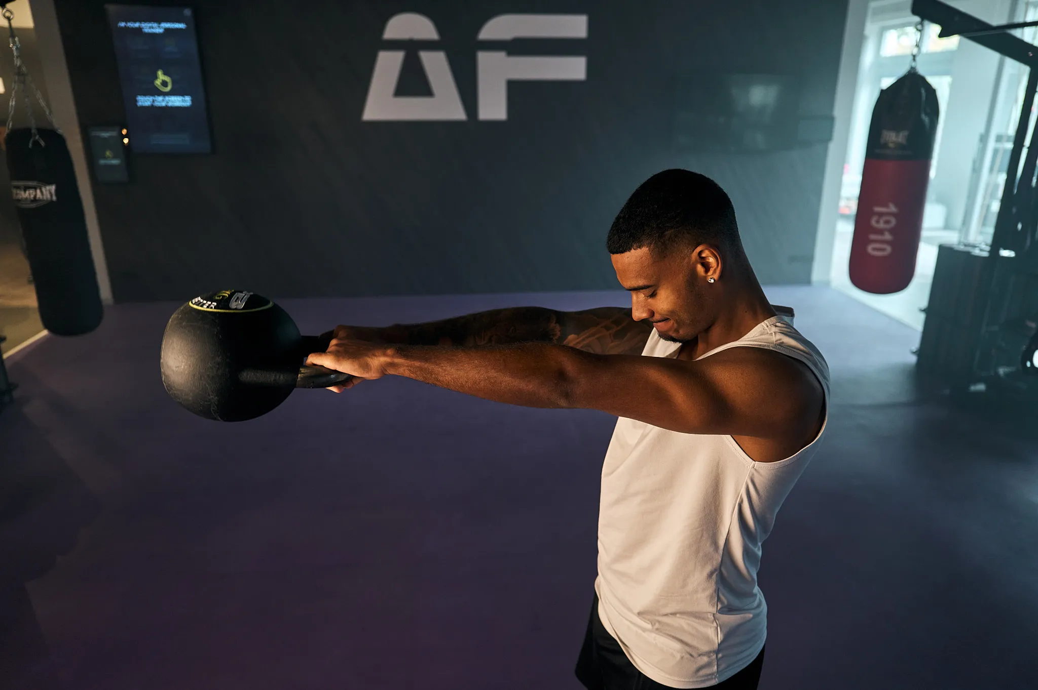 Mann in weißem Tanktop hält in einem Fitnessstudio eine Kettlebell mit ausgestreckten Armen.