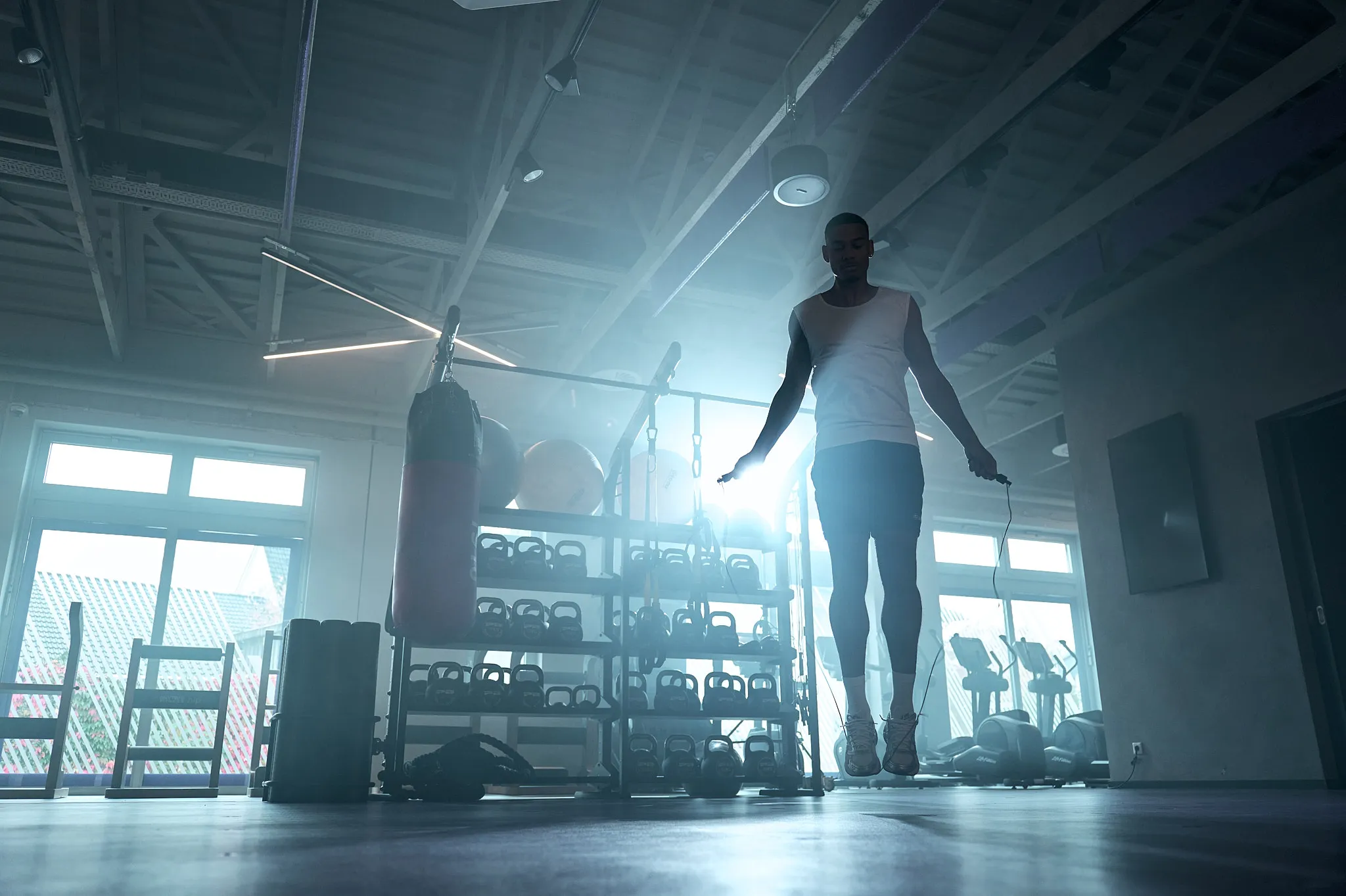 Mann beim Seilspringen während des Trainings in einem Fitnessstudio mit Kettlebells und Cardiogeräten im Hintergrund.