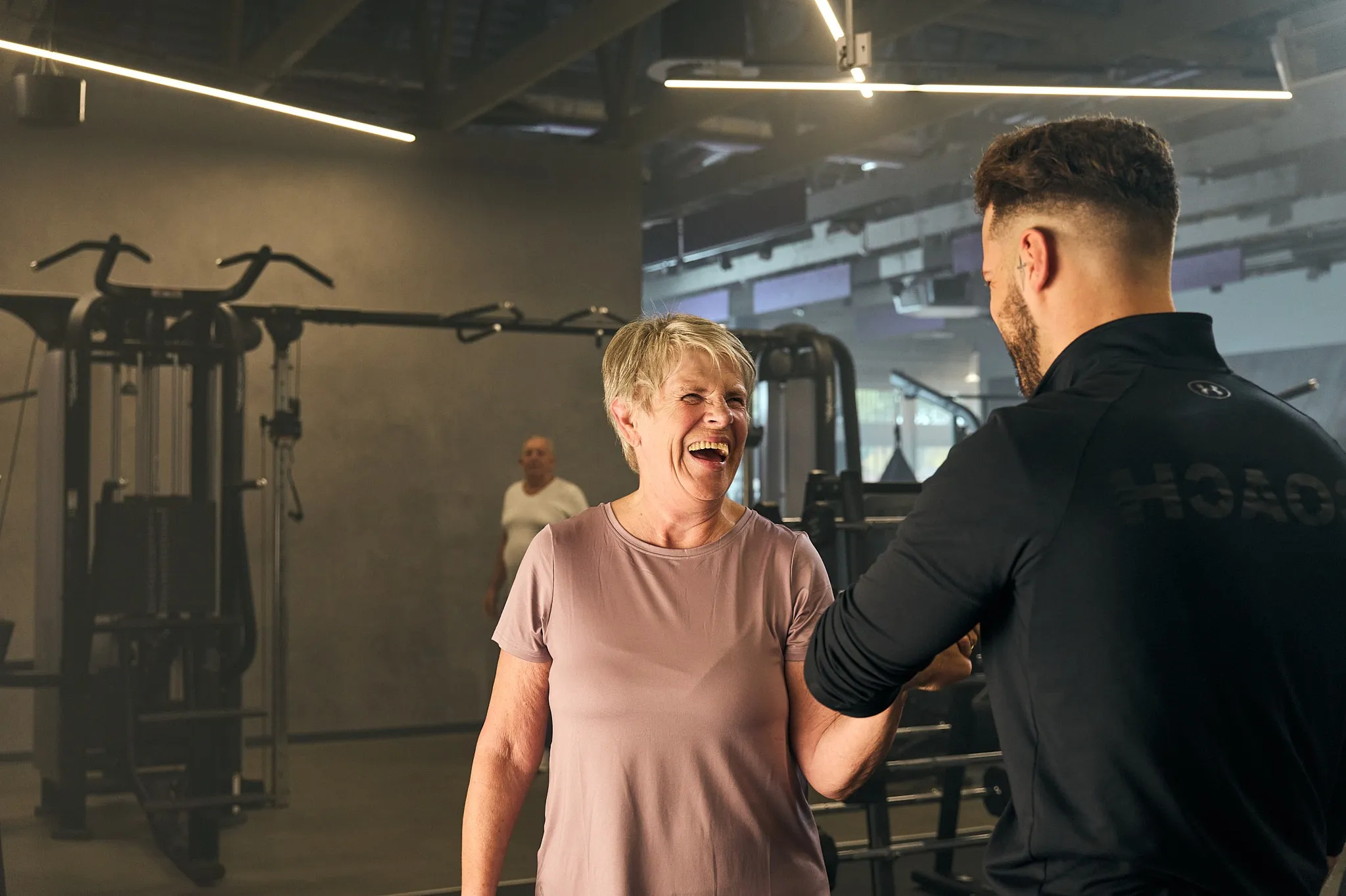 Fitness coach and a smiling older woman bumping fists in a gym with exercise equipment in background.