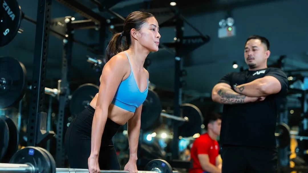 Woman executing a barbell lift while monitored by an Anytime Fitness personal trainer.