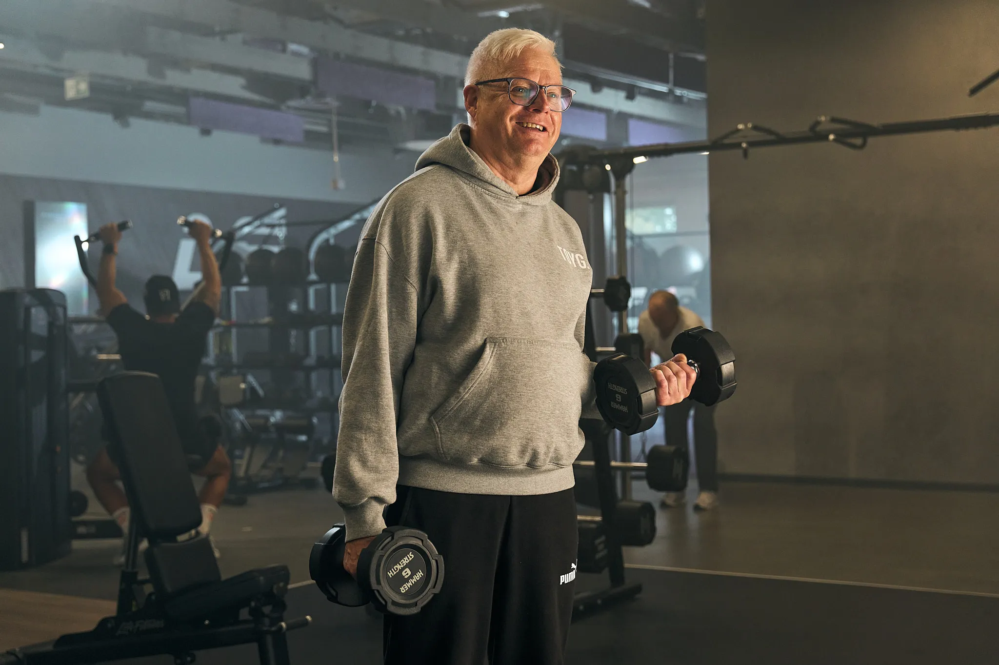 Smiling older man lifting dumbbells in a gym with other people exercising in the background.