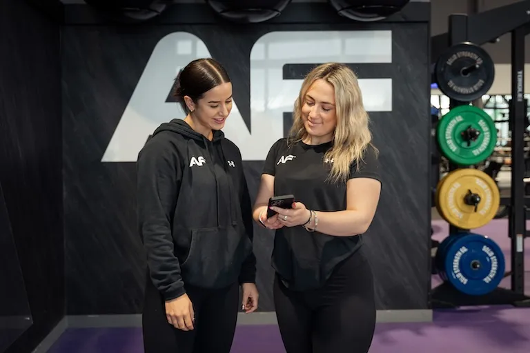 Two women in AF workout clothes at gym, one showing the other something on her phone.