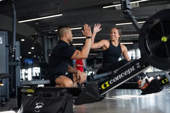 Man and woman in gym giving each other a high-five while sitting on rowing machines.
