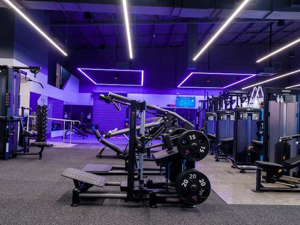 Modern gym area with various workout machines, weights, and purple neon lights on the ceiling.