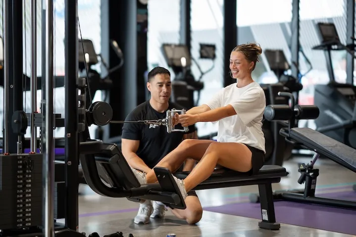 Woman exercising on a rowing machine with a trainer watching her in a gym.