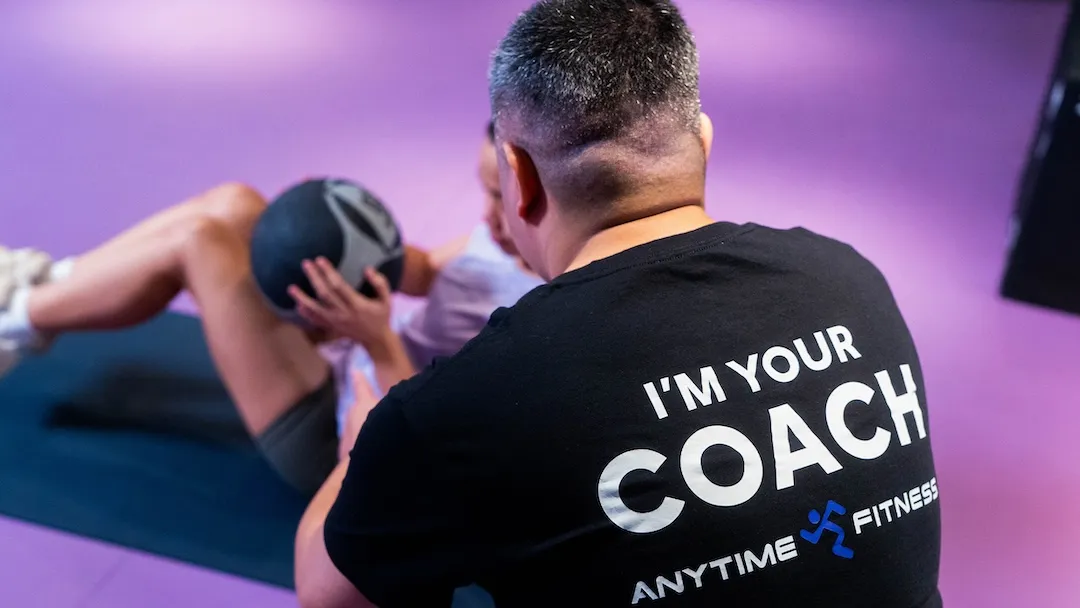 Anytime Fitness personal trainer coaching a client performing a medicine ball core exercise.