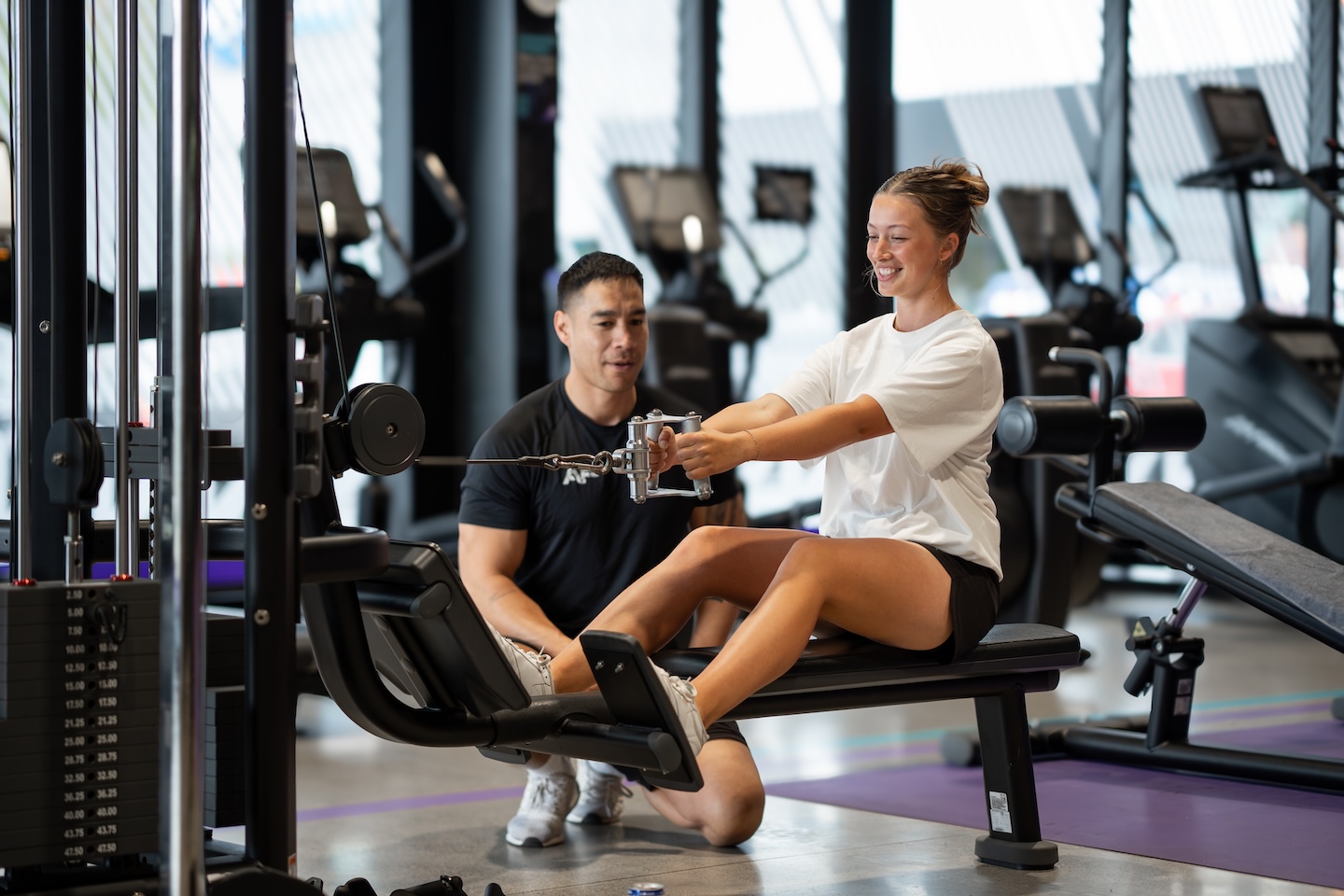 A personal trainer assists a man lifting heavy dumbbells on a bench in a busy gym with various equipment in the background.