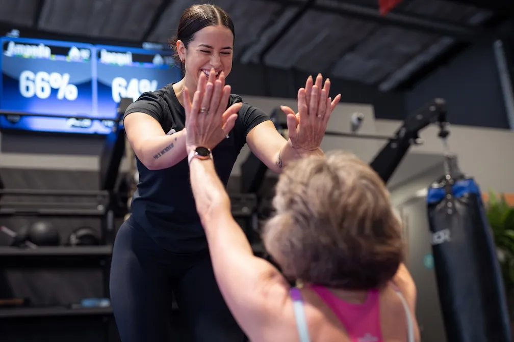 Trainer high fiving an older member with both hands in a gym, heart rate stats displayed on a screen behind them