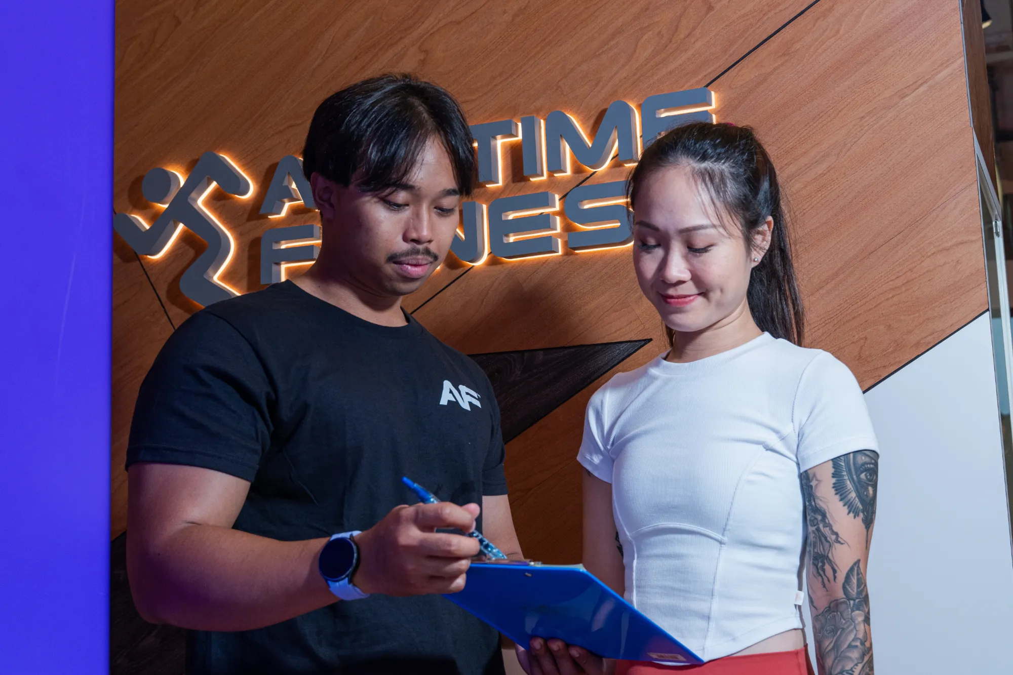 Man in black shirt with smartwatch shows clipboard to smiling tattooed woman in white at Anytime Fitness.